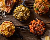 Four varieties of mac and cheese on a wooden table including chicken mac and cheese, meatball mac and cheese and broccoli mac and cheese